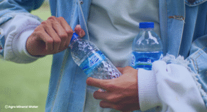 Tangan seseorang sedang membuka botol air mineral AGRO, dengan satu botol lain dipegang di tangan satunya, menampilkan kemasan produk sebagai simbol hidrasi yang sehat.