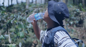 Seorang pria menggunakan topi dan ransel sedang meminum Agro Mineral Water di tengah suasana alam terbuka, menekankan pentingnya hidrasi selama aktivitas fisik di luar ruangan.