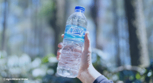 Tangan memegang botol air mineral AGRO di luar ruangan dengan latar belakang pepohonan.