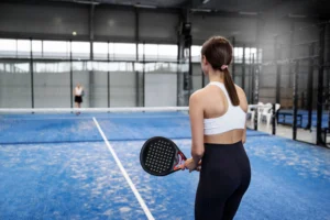 Seorang wanita bermain padel di lapangan indoor biru dengan raket padel hitam, menggambarkan olahraga padel yang semakin populer di dunia.