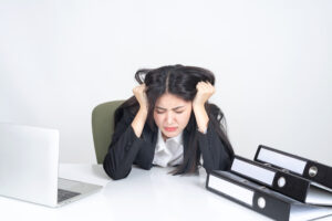 Wanita karier mengenakan setelan jas hitam tampak stres dan menarik rambutnya di meja kerja dengan laptop dan tumpukan dokumen. Ilustrasi burnout akibat beban kerja berlebihan di kantor.