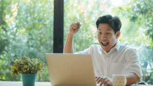 Pria muda tersenyum lebar dan mengangkat tangan dengan ekspresi gembira di depan laptop, suasana kerja penuh semangat dengan latar belakang jendela hijau. Ilustrasi motivasi, produktivitas, dan keberhasilan.